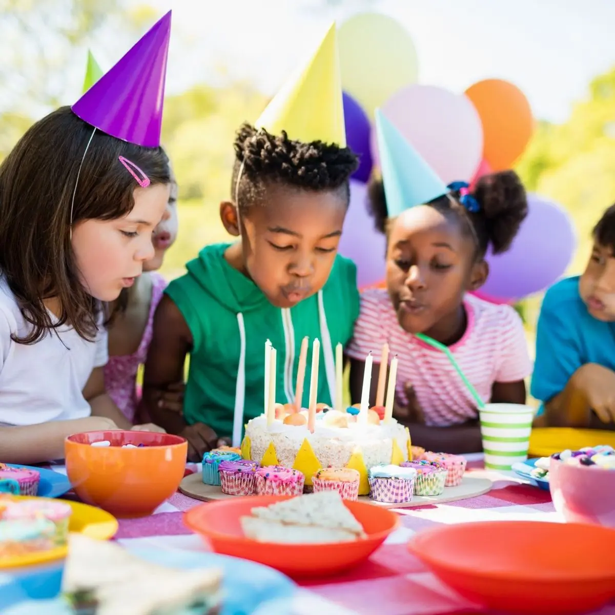 Des enfants portant des chapeaux de fête colorés soufflent les bougies d'un gâteau d'anniversaire lors d'une fête en plein air. La table est décorée avec des cupcakes, des assiettes colorées et des ballons en arrière-plan.